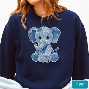 May include: A navy blue crewneck sweatshirt featuring a central graphic of a baby elephant. The elephant is designed with a patchwork pattern in various shades of blue, resembling denim fabric textures with visible stitching along the edges.