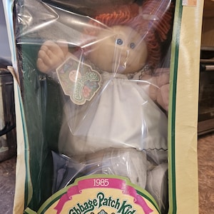 May include: A vintage Cabbage Patch Kids doll in its original packaging. The doll has red yarn hair, blue eyes, and is wearing a white dress. The packaging features the Cabbage Patch Kids logo and the year 1985. The text "I'm unique. I'm adoptable!" is also visible.