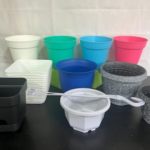 May include: Assortment of plastic plant pots in various colors, including white, blue, green, pink, and black. The pots are arranged in a group on a table.