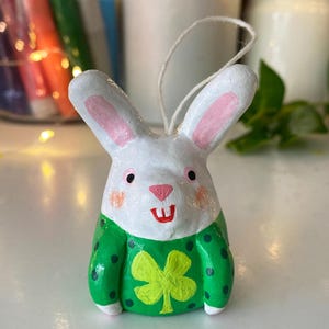 May include: A handmade white bunny ornament with pink inner ears, rosy cheeks, and a red mouth. The bunny wears a green sweater with a yellow shamrock. The ornament has a string for hanging. The text "cute attack" is on a pink sticker.
