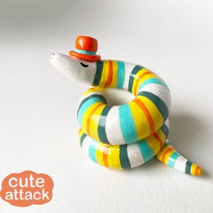 May include: A whimsical, colorful ceramic snake figurine with a small orange top hat. The snake is coiled, featuring stripes of yellow, blue, orange, and white. The text "cute attack" is in a rounded orange box.