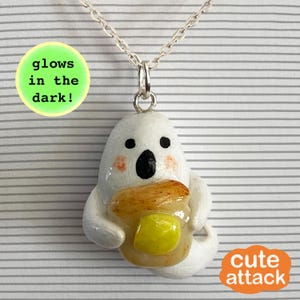 May include: A silver-toned chain necklace featuring a white ghost pendant holding a piece of toast with a yellow butter pat. The ghost has black eyes, a black mouth, and pink cheeks. A green circular label reads glows in the dark! and an orange label reads cute attack.