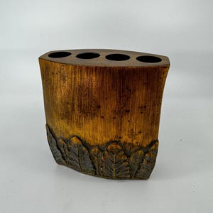 May include: A brown wooden toothbrush holder with four slots and a carved leaf design around the bottom.