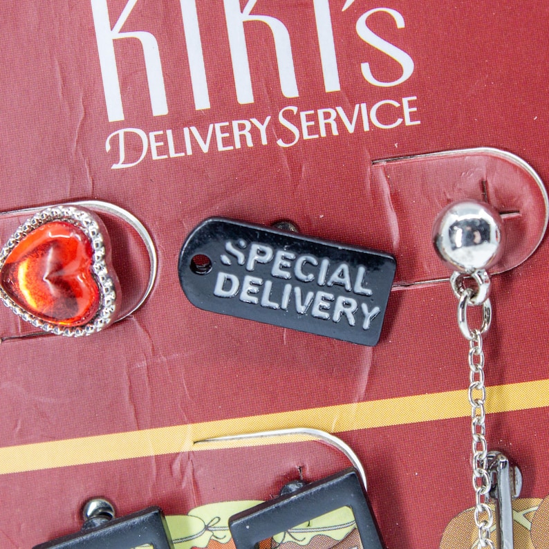 May include: Close-up of a display featuring earrings and a pin. The pin is heart-shaped with a red center and silver trim. A black tag reads "SPECIAL DELIVERY". The display background is red with the text "Kiki's Delivery Service".