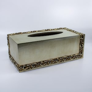 May include: A rectangular tissue box with a light green finish and a decorative gold-colored border. The box has an oval opening on top for tissue dispensing. The gold border features an ornate, swirling design.