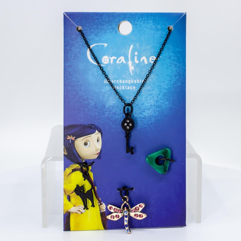 May include: An interchangeable necklace set featuring a black chain, a key pendant, a green triangle pendant, and a dragonfly pendant with pink and blue accents. The backing card has the word "Coraline" and an illustration of a character.