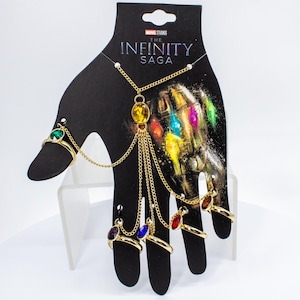 May include: A gold-colored hand chain and ring set featuring colorful gemstones. The set includes rings for each finger and a chain that connects to a necklace. The packaging features the text "Marvel Studios The Infinity Saga".