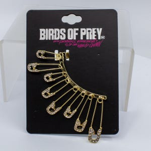 May include: Gold-toned ear cuff featuring safety pin design with clear rhinestones. The cuff is displayed on a black card with the text "BIRDS OF PREY" and "HARLEY QUINN".