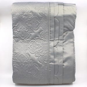 May include: A silver-gray quilted blanket with an ornate, repeating floral pattern on one side and vertical channel stitching on the other. The blanket has a satin-like sheen, suggesting a luxurious feel. The overall design is elegant and suitable for a bedroom.