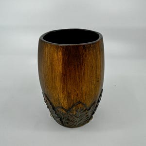 May include: A brown ceramic vase with a leaf design around the bottom. The vase has a smooth, wood-like finish.