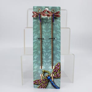 May include: Two decorative hair sticks with dragonfly designs. The dragonflies have red and purple jeweled wings, blue bodies, and gold-colored metal stems. They are displayed on a teal card with a dragonfly pattern.