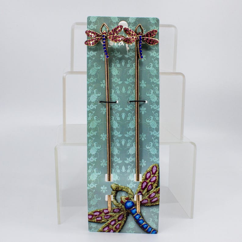 May include: Two decorative hair sticks with dragonfly designs. The dragonflies have red and purple jeweled wings, blue bodies, and gold-colored metal stems. They are displayed on a teal card with a dragonfly pattern.
