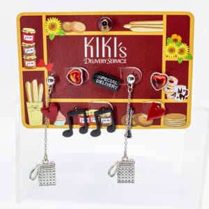 May include: A pair of earrings with a Kiki's Delivery Service theme. One earring features a silver camera charm with a chain, a red heart, and a musical note. The other earring has a silver camera charm, a black tag that says "Special Delivery", and a heart-shaped charm.