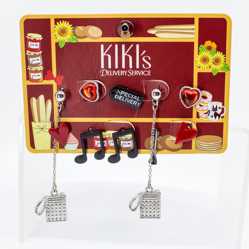 May include: A pair of earrings with a Kiki's Delivery Service theme. One earring features a silver camera charm with a chain, a red heart, and a musical note. The other earring has a silver camera charm, a black tag that says "Special Delivery", and a heart-shaped charm.