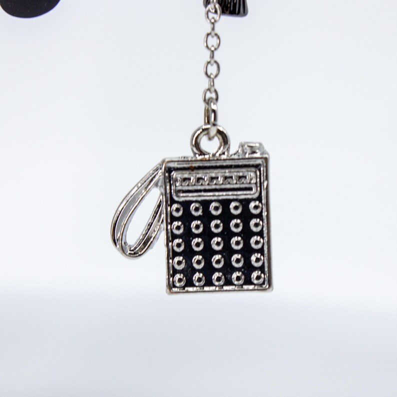 May include: A silver-colored charm in the shape of a vintage calculator, with a black keypad and a small handle. The charm is suspended from a delicate chain, perfect for jewelry or accessories.