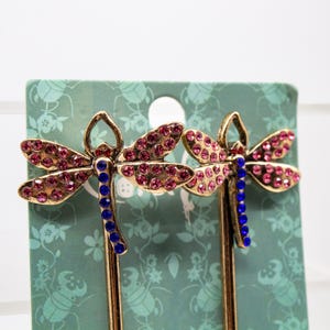 May include: Two dragonfly hair pins. The pins have gold-colored metal bodies, with pink rhinestone wings and blue rhinestone bodies. The pins are displayed on a green floral patterned card.