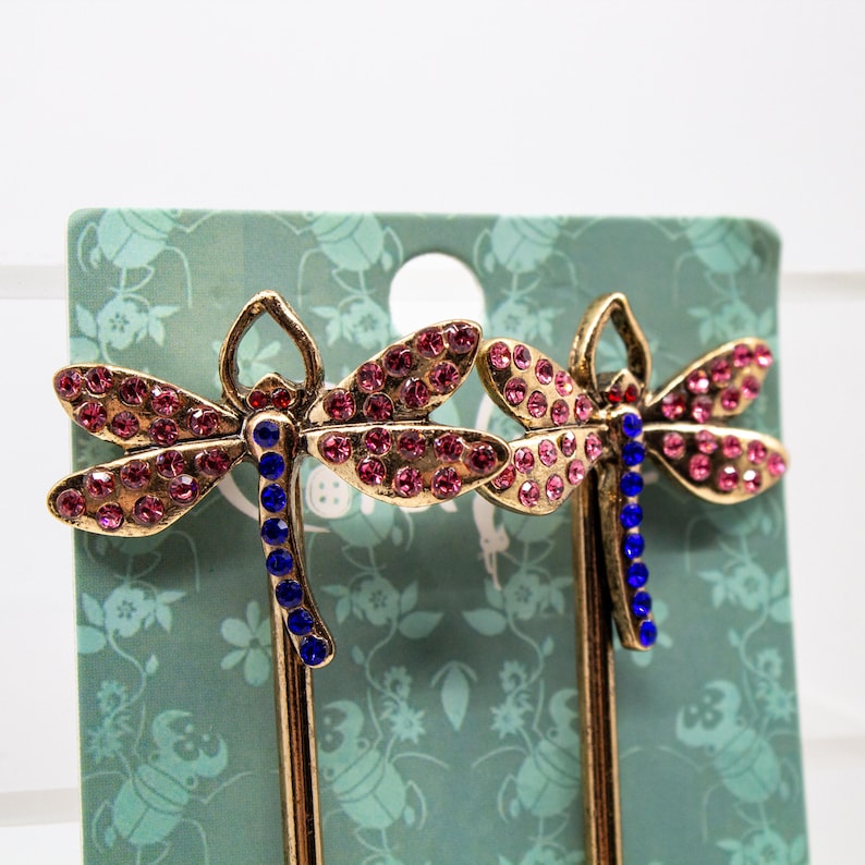 May include: Two dragonfly hair pins. The pins have gold-colored metal bodies, with pink rhinestone wings and blue rhinestone bodies. The pins are displayed on a green floral patterned card.