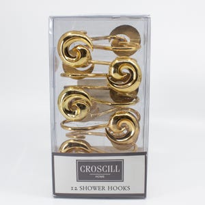 May include: A clear plastic box containing twelve gold-colored shower hooks. Each hook features a decorative swirl design. The box has a label that reads "Croscill Home" and "12 Shower Hooks".
