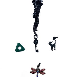 May include: A collection of charms and pendants on a white background. Includes a black key pendant, a green triangle, a black cat charm, and a dragonfly pendant with pink and blue accents.