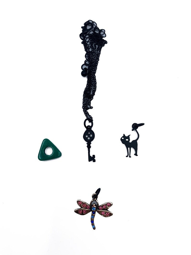 May include: A collection of charms and pendants on a white background. Includes a black key pendant, a green triangle, a black cat charm, and a dragonfly pendant with pink and blue accents.