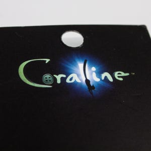 May include: Black tag with a circular hole at the top. The word "Coraline" is written in a stylized, green font with a button-like design for the letter "O". A blue and white light effect is behind the word.