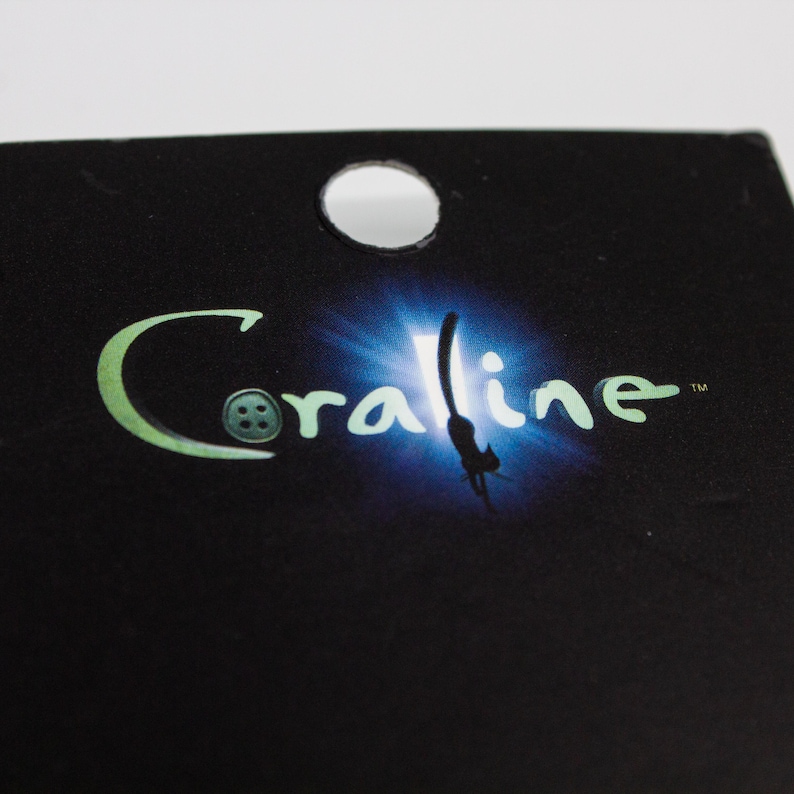 May include: Black tag with a circular hole at the top. The word "Coraline" is written in a stylized, green font with a button-like design for the letter "O". A blue and white light effect is behind the word.