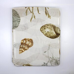 May include: A folded rectangular fabric featuring a seashell design. The fabric has a white background with a wave pattern and illustrations of seashells in shades of brown, tan, and green. Seaweed-like elements are also present.