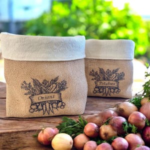 Puede incluir: Dos bolsas de almacenamiento de arpillera con la parte superior de color crema. Una bolsa tiene la palabra "Onions" y la otra "Potatoes" impresa en la parte delantera. Las bolsas están sobre una superficie de madera con cebollas frescas y vegetación.