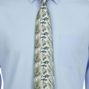 May include: A necktie featuring a repeating pattern of blue fish and green seaweed on a light cream background, worn over a light blue button-down shirt with a pointed collar and a single chest pocket on the left side of the garment.