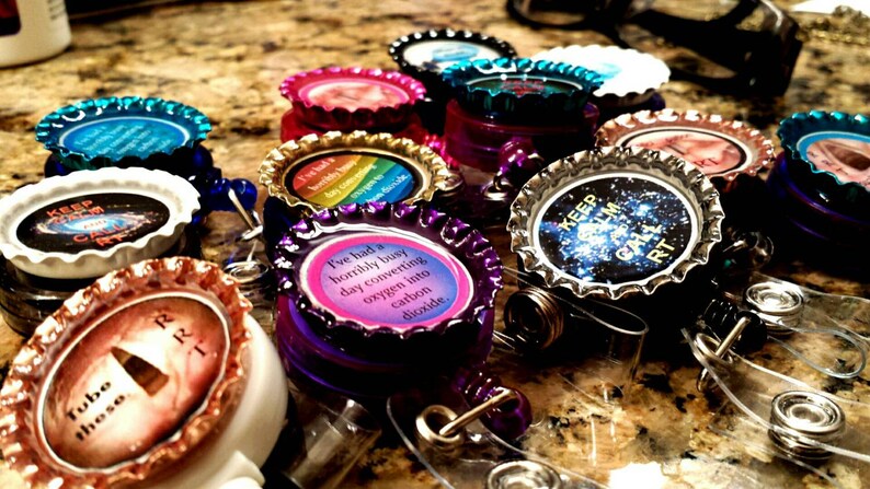 May include: A collection of colorful bottle cap badge reels with various sayings, including "Keep Calm and Carry On", "Tube Those", and "I've had a horribly busy day converting oxygen into carbon dioxide".