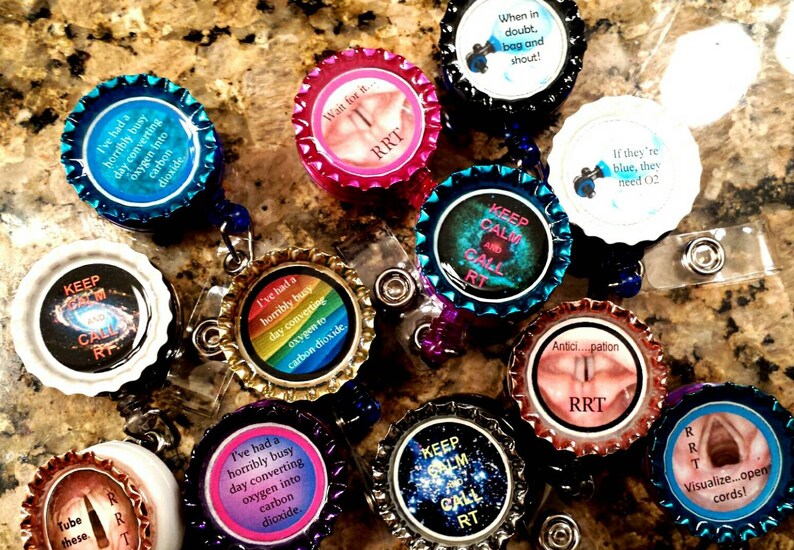 May include: A collection of colorful, round, plastic badge holders with various respiratory therapy-related sayings and images. The sayings include "Keep Calm and Call RT", "If they're blue, they need O2", "I've had a horribly busy day converting oxygen into carbon dioxide", and "Wait for it... RRT".
