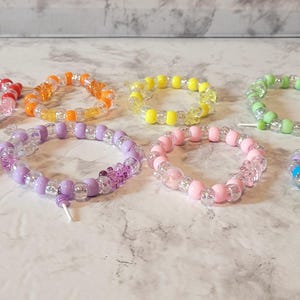 May include: A collection of eight beaded bracelets in various colors, including red, orange, yellow, green, blue, purple, pink, and rainbow. Each bracelet features a mix of clear and colored beads, strung together with a white string.