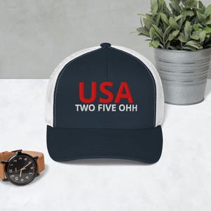 Puede incluir: Gorra de camionero azul marino y blanca con la palabra "USA" en letras rojas en bloque sobre el texto "TWO FIVE OHH" en blanco. La gorra tiene una visera curva y una parte trasera de malla.