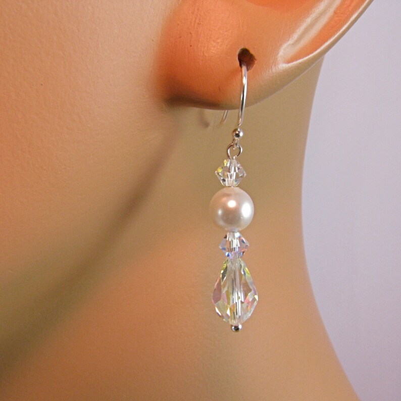 May include: Dangle earrings featuring a white pearl and clear, faceted crystals. The earrings have a silver-colored hook and are designed to be worn in pierced ears. The crystals reflect light, creating a sparkling effect.