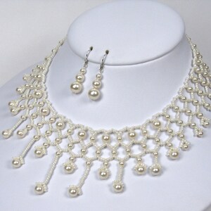 Op de afbeelding: Een cr&egrave;me-kleurige kralenketting en bijpassende oorbellen set. De ketting heeft een delicaat, open ontwerp met bungelende parelaccenten. De oorbellen hebben elk drie parels. De sieraden worden gepresenteerd op een witte mannequin hoofd.