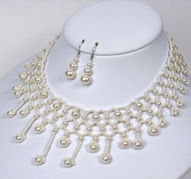 Op de afbeelding: Een cr&egrave;me-kleurige kralenketting en bijpassende oorbellen set. De ketting heeft een delicaat, open ontwerp met bungelende parelaccenten. De oorbellen hebben elk drie parels. De sieraden worden gepresenteerd op een witte mannequin hoofd.