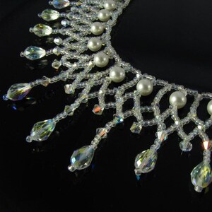 May include: A close-up of a clear, beaded necklace with iridescent crystal accents and faux pearl beads. The necklace has a delicate, web-like design with teardrop-shaped crystal pendants hanging from the bottom.