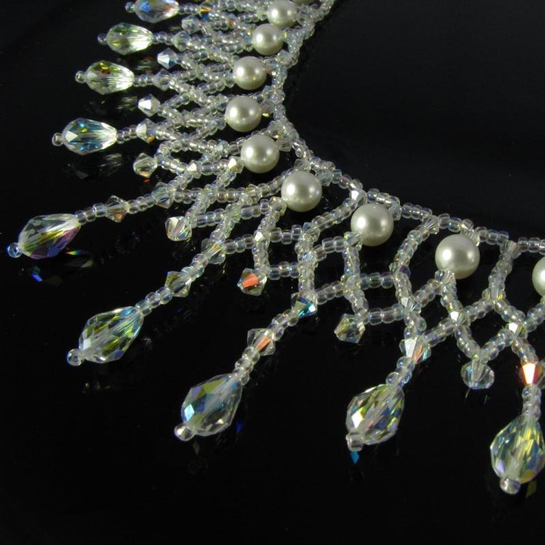 May include: A close-up of a clear, beaded necklace with iridescent crystal accents and faux pearl beads. The necklace has a delicate, web-like design with teardrop-shaped crystal pendants hanging from the bottom.