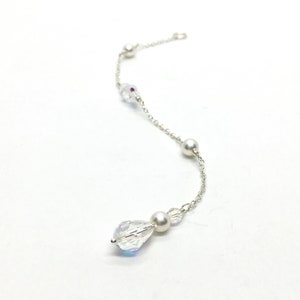 May include: A delicate silver chain adorned with clear, iridescent crystals, small pearls, and a larger teardrop-shaped crystal. The jewelry piece is displayed against a white background, showcasing its elegant design.