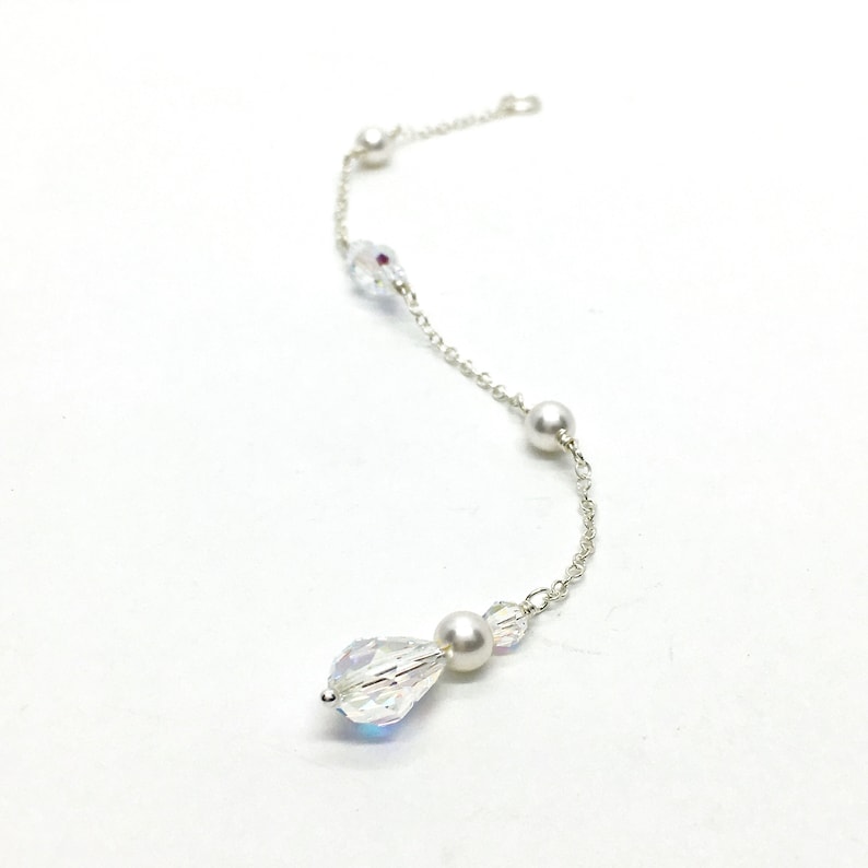 May include: A delicate silver chain adorned with clear, iridescent crystals, small pearls, and a larger teardrop-shaped crystal. The jewelry piece is displayed against a white background, showcasing its elegant design.
