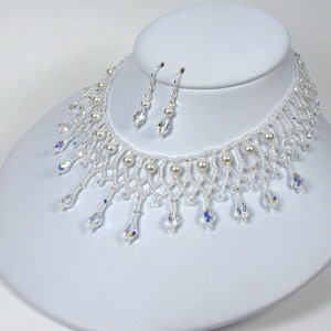 May include: A delicate white beaded necklace and matching earrings set. The necklace features faux pearls and teardrop-shaped iridescent crystals. The earrings have a similar design with pearls and crystals. The jewelry is displayed on a white mannequin bust.