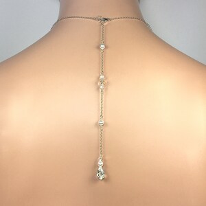 May include: A silver-toned necklace with a delicate chain and a drop pendant. The necklace features small pearls and clear crystals along the chain, with a teardrop-shaped crystal at the end. The necklace is designed to be worn on the back.