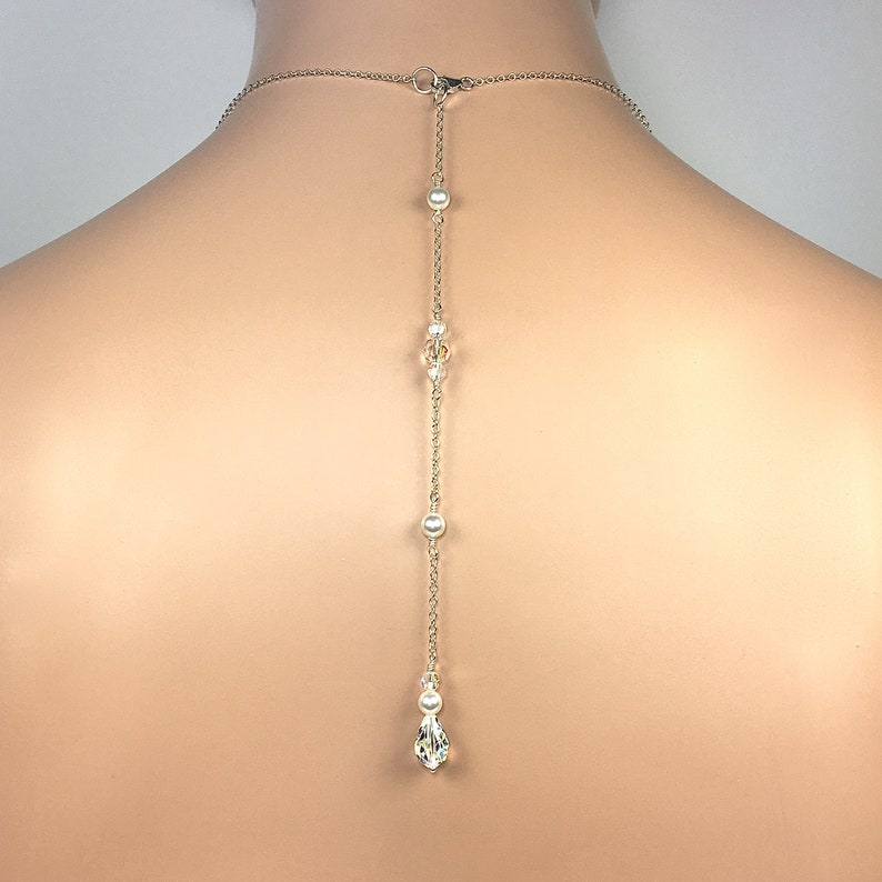 May include: A silver-toned necklace with a delicate chain and a drop pendant. The necklace features small pearls and clear crystals along the chain, with a teardrop-shaped crystal at the end. The necklace is designed to be worn on the back.