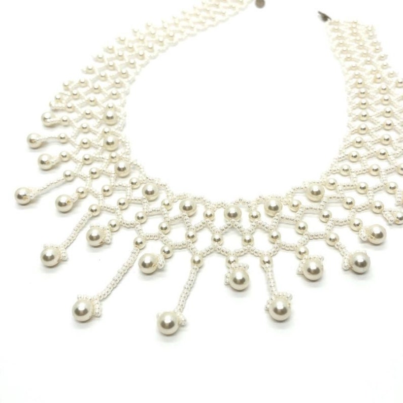 Op de afbeelding: Een delicate, cr&egrave;mekleurige parelketting. De ketting heeft een netvormig ontwerp met bungelende parelaccenten. De parels zijn rond en gelijkmatig verdeeld, wat een klassieke en elegante uitstraling cre&euml;ert. De achtergrond is wit.
