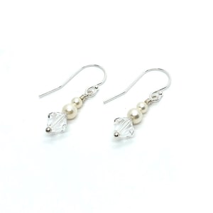 May include: A pair of dangle earrings with a silver hook. Each earring features a clear, square-cut crystal, two small pearls, and a larger pearl. The earrings are set against a white background.