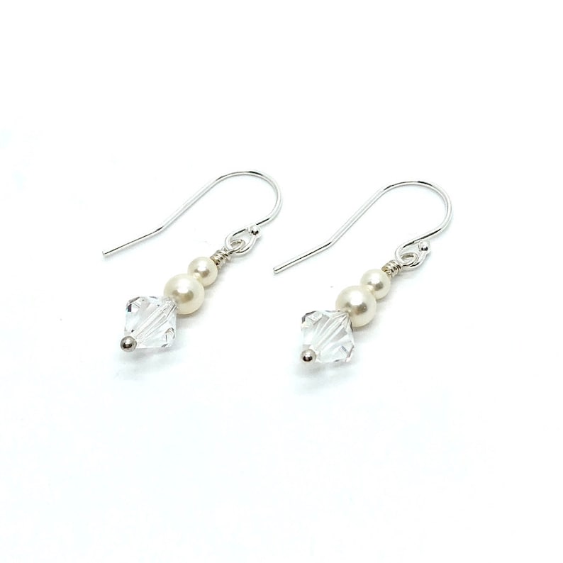 May include: A pair of dangle earrings with a silver hook. Each earring features a clear, square-cut crystal, two small pearls, and a larger pearl. The earrings are set against a white background.