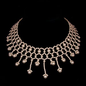 Op de afbeelding: Een delicate parelketting met een geometrisch ontwerp. De ketting heeft een reeks onderling verbonden parelsnoeren, waardoor een webachtig patroon ontstaat. Verschillende strengen hangen naar beneden, elk eindigend met een grotere parel. De ketting staat tegen een zwarte achtergrond.