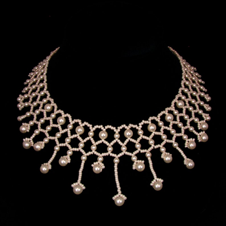 Op de afbeelding: Een delicate parelketting met een geometrisch ontwerp. De ketting heeft een reeks onderling verbonden parelsnoeren, waardoor een webachtig patroon ontstaat. Verschillende strengen hangen naar beneden, elk eindigend met een grotere parel. De ketting staat tegen een zwarte achtergrond.