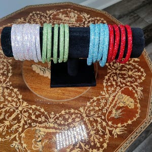 May include: A display of sparkling bangles in various colors. The bangles are arranged on a black velvet stand. Colors include iridescent, light green, light blue, and red. The bangles are displayed on a decorative wooden table.