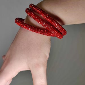 May include: Three red, sparkly bracelets are stacked on a wrist. The bracelets are covered in small, glittering red stones. The background is a solid, neutral gray.